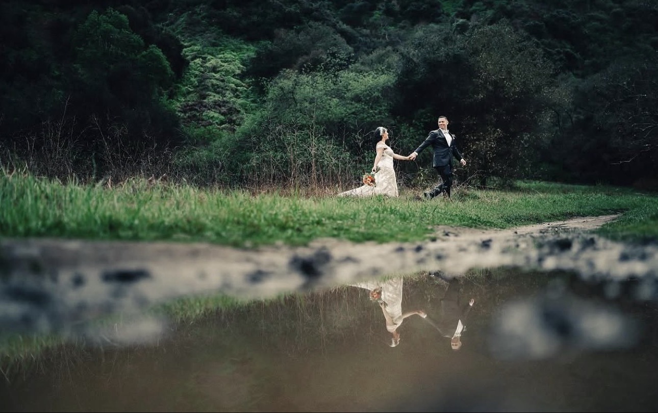 Anthony and Elizabeth walking hand in hand through tall grass on their wedding day, reflected in the water below